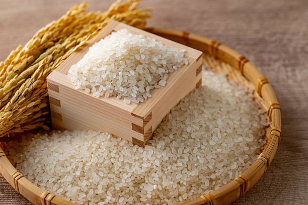 A close-up of Japanese white rice in a traditional wooden masu box, with golden rice stalks in the background. This image captures the freshness and tradition of Japanese agriculture.